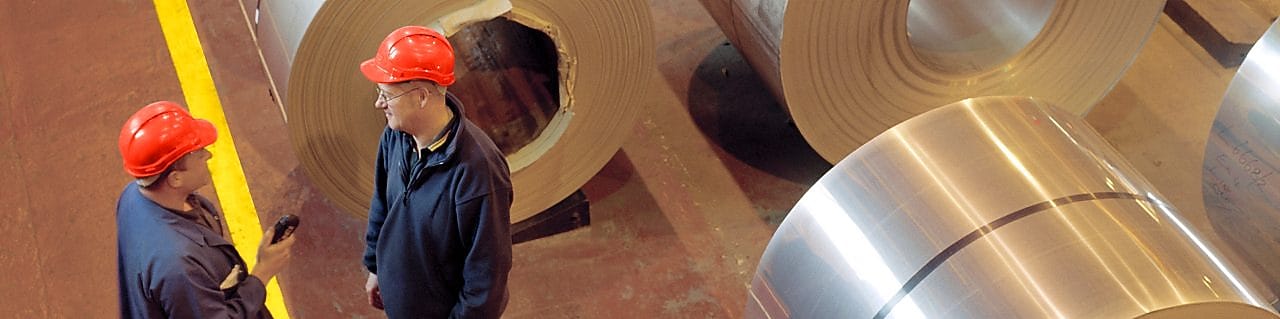 Two factory workers talking next to large rolls of steel