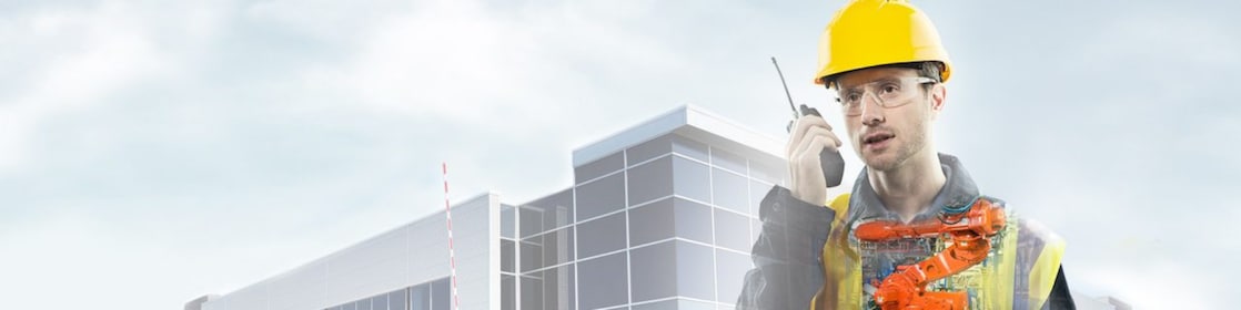 노란색 안전모를 쓰고 무전기를 들고 있는 남자가 큰 건물을 배경으로 현장 앞에 서 있습니다.